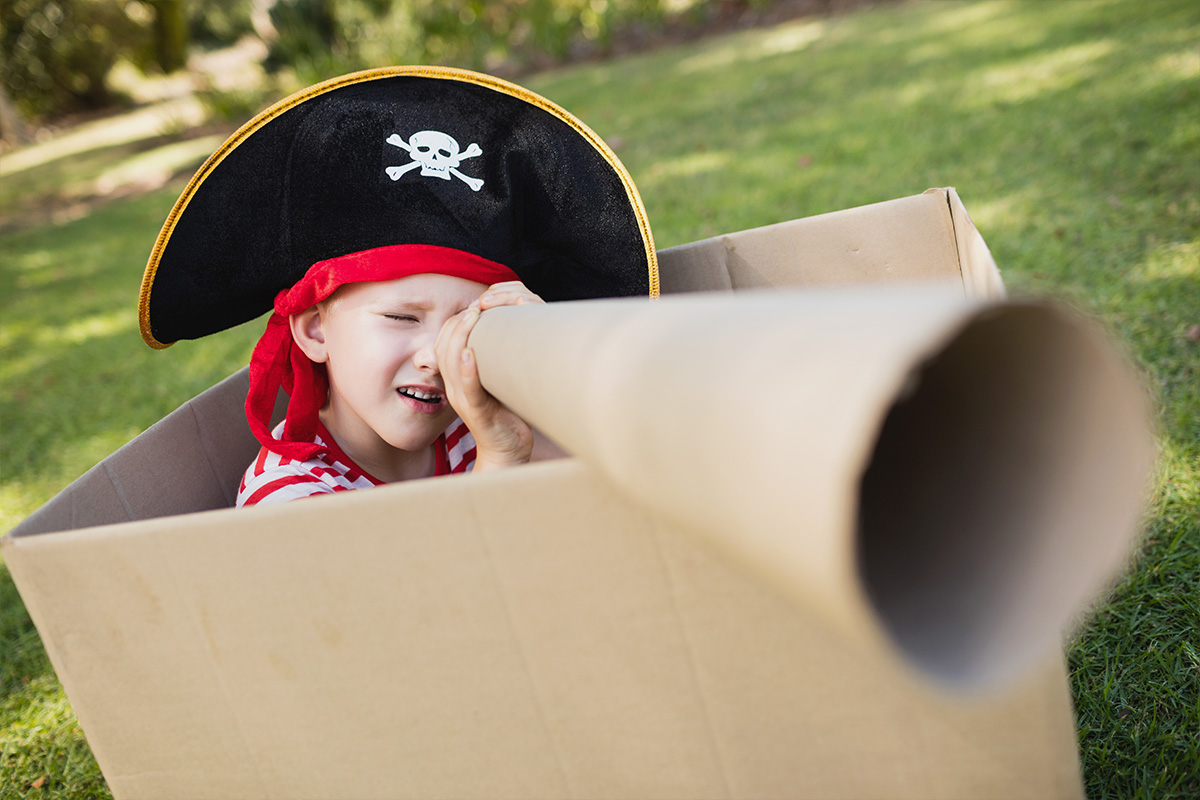 Bambino vestito da pirata che gioca dentro una scatola di cartone e una un tubo di cartone come binocolo